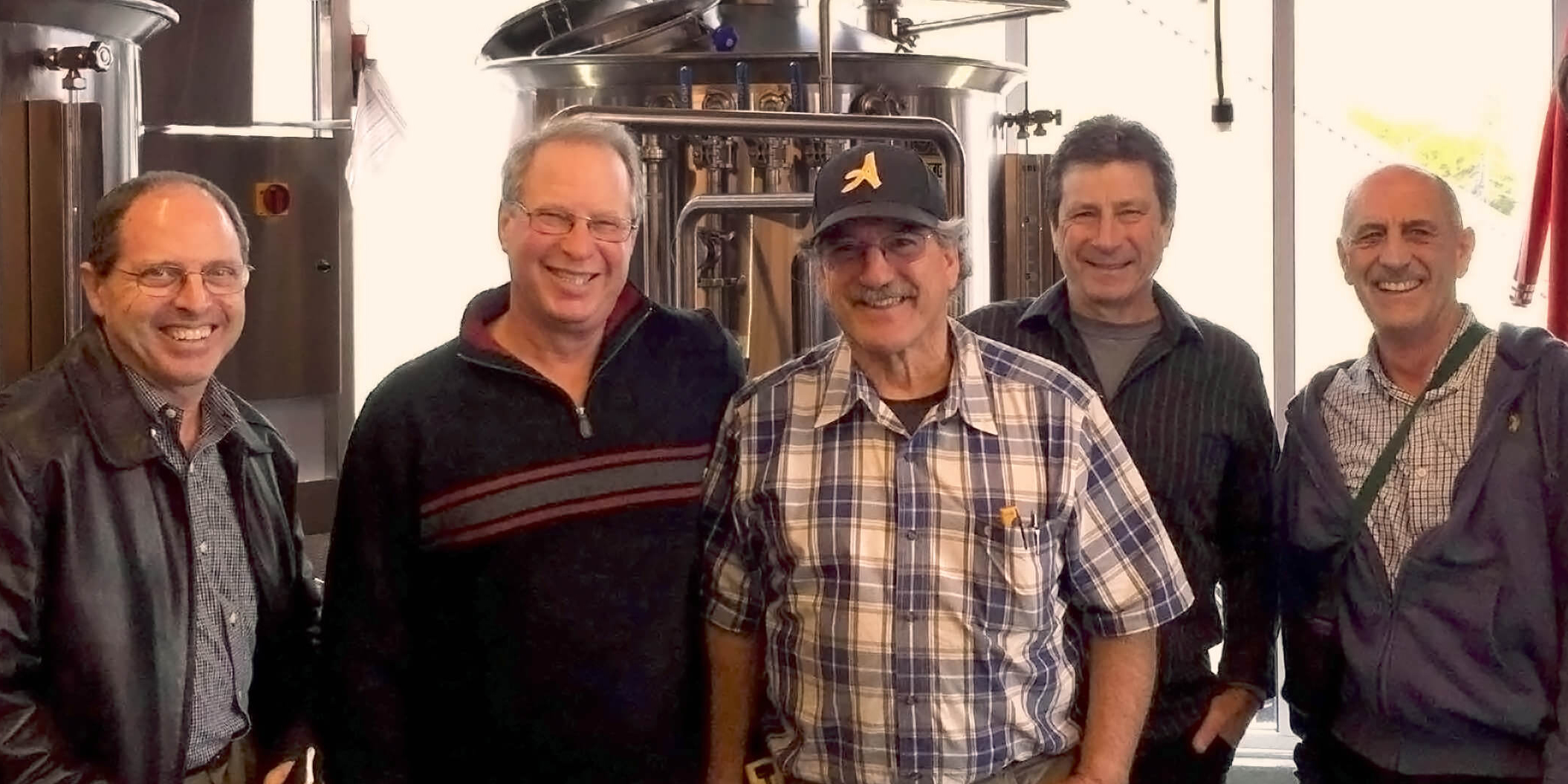 Five smiling men — Sutti Associates team members — standing in front of large metal brewery tanks.